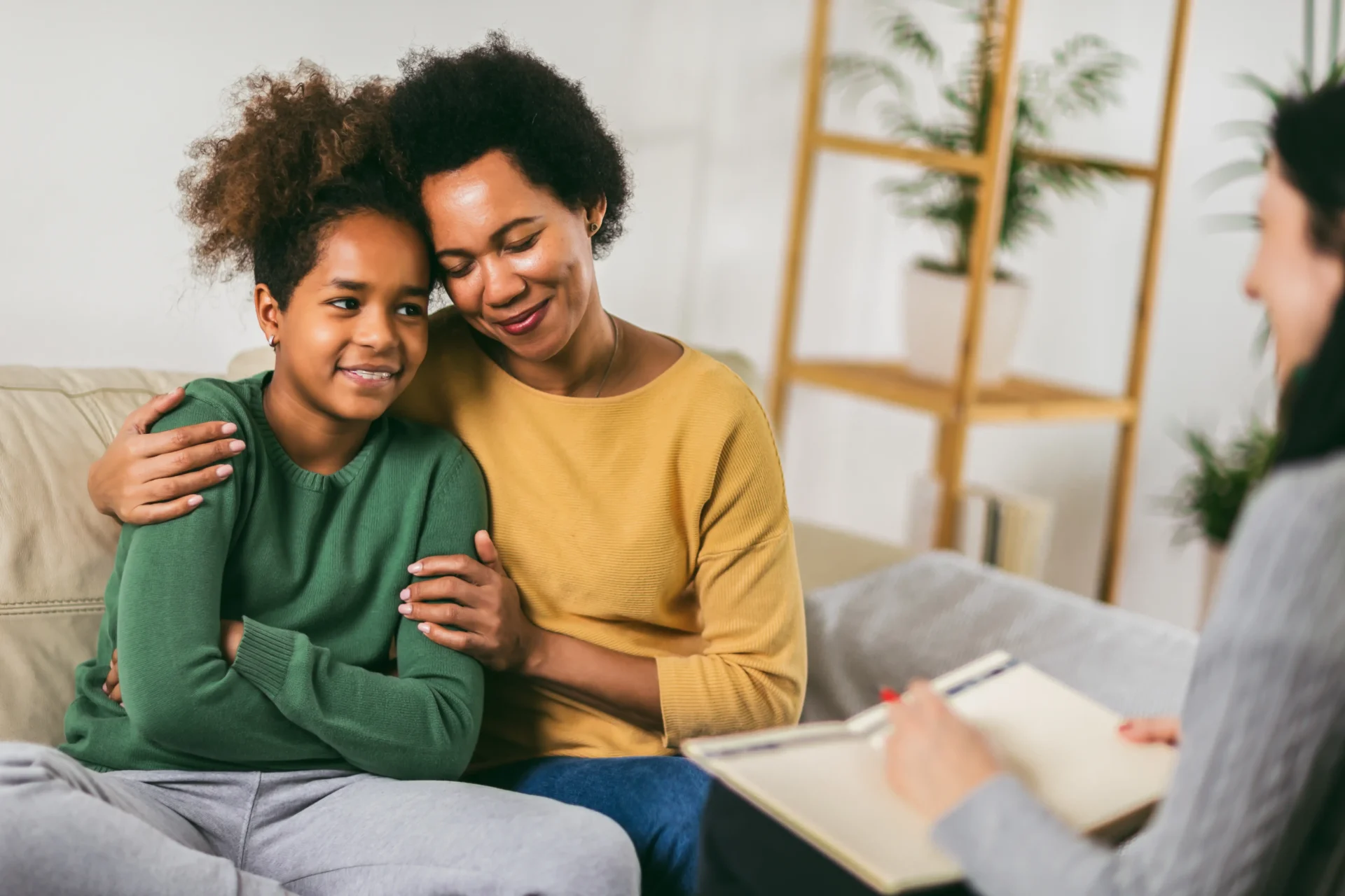 A woman in a yellow sweater comforts a girl in a green sweater, both sitting on a couch during a counseling session.
