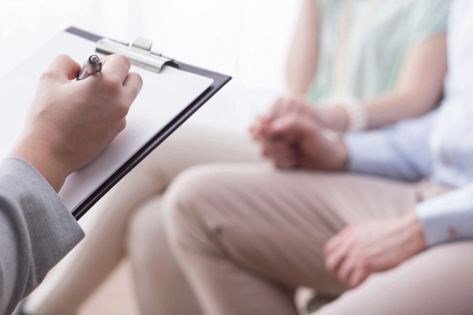 A person writes on a clipboard while two individuals hold hands in a supportive, calm atmosphere during a counseling session.