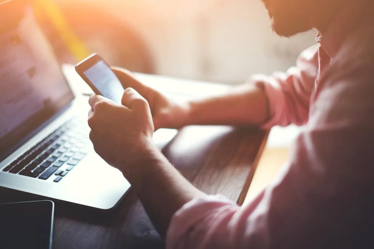 Man sitting at home, using a smartphone in one hand and a laptop on a table in front of him.